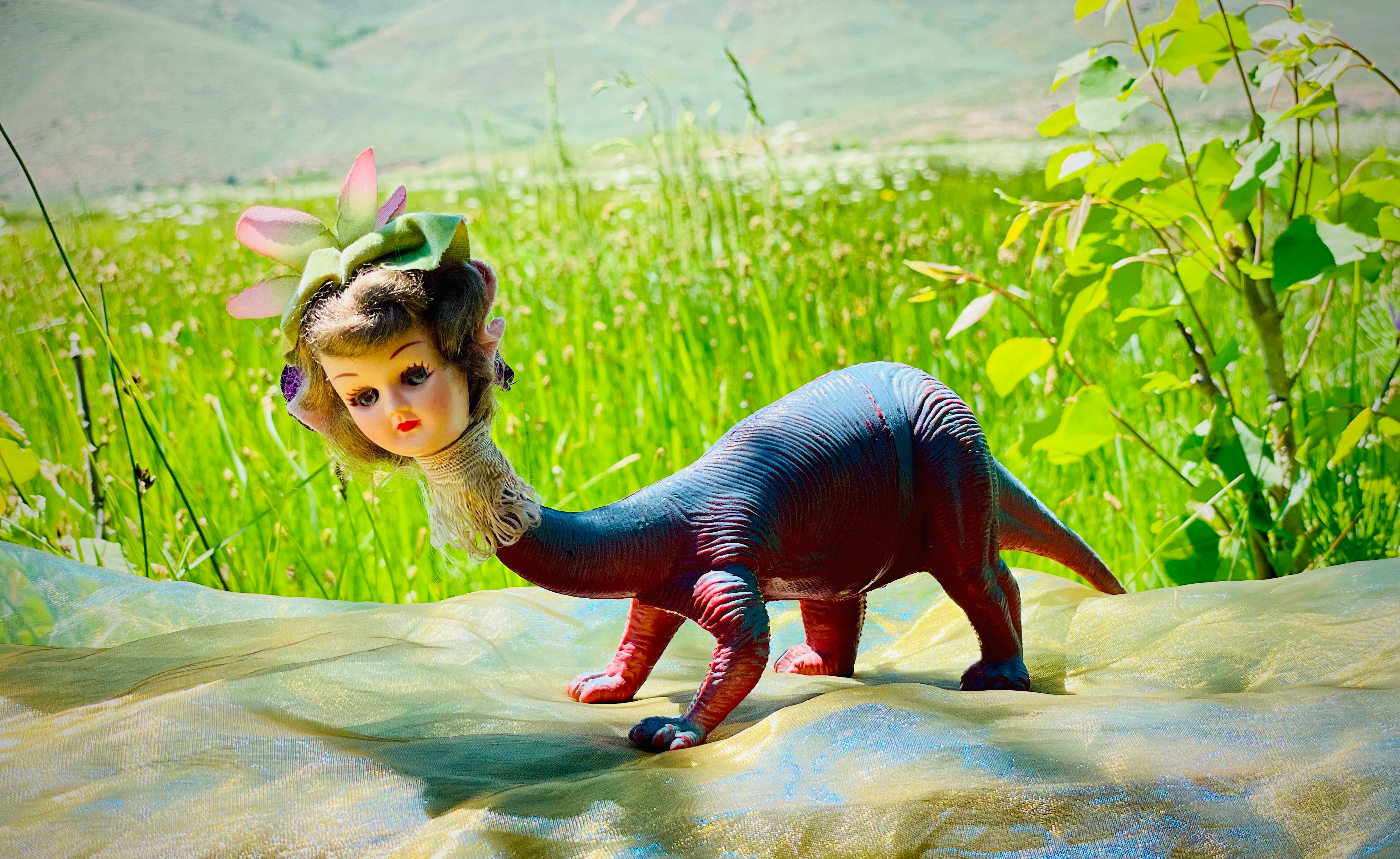 Vintage dinosaur body with vintage doll head, felt hat and flower, ornate neck trim. 7" long 3" tall 2" wide. Displayed on gold gauze. Green grass in the background