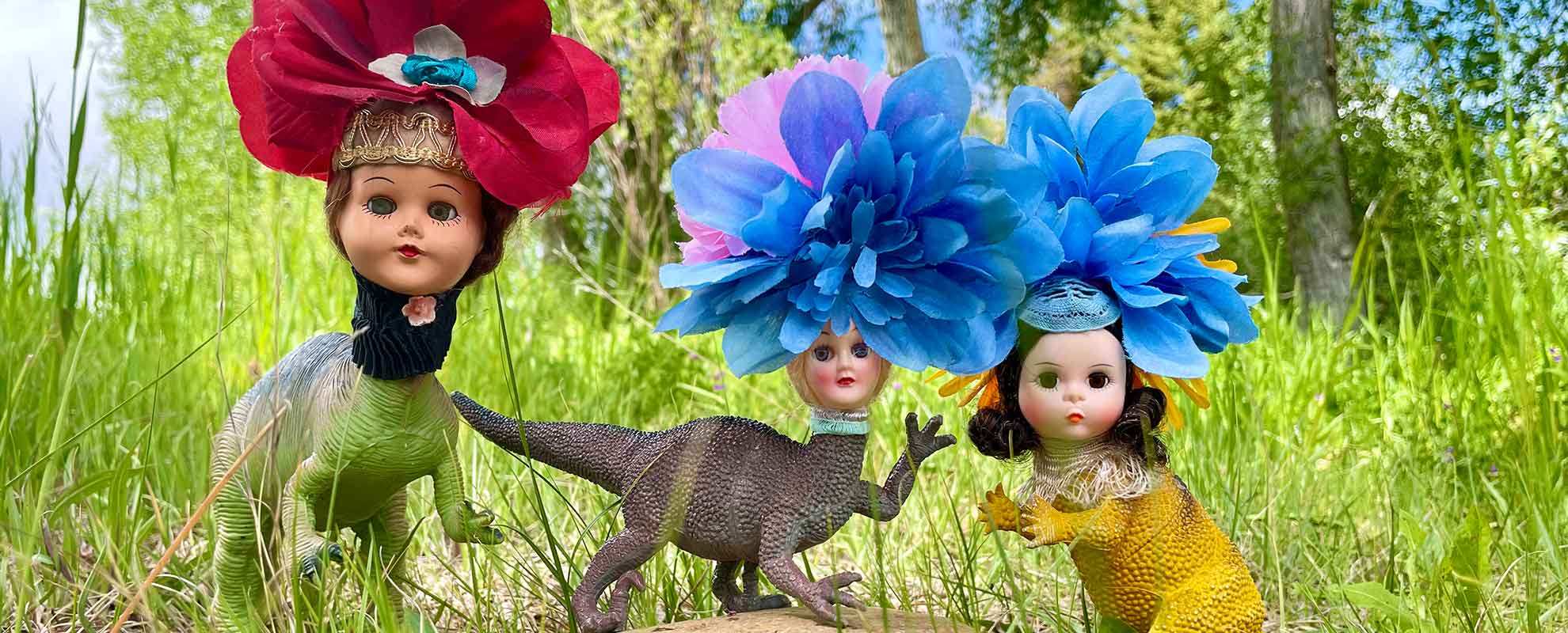 Three art dolls in a gassy field with woman doll heads with big flower headdresses and dinosaur bodies